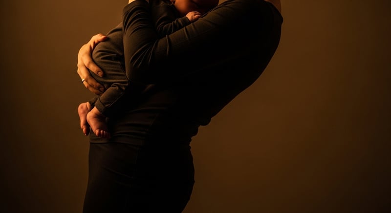 Editorial side photograph of a parent cradling an infant low in front, upper back rounded and torso leaning back to counterbalance, warm tones, face cropped above the hairline