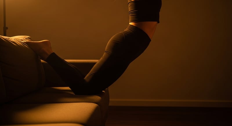 Editorial side-profile photograph of a person setting up the couch stretch, back shin rising up a padded couch edge, front foot planted, torso upright, warm amber light, face cropped above the nose