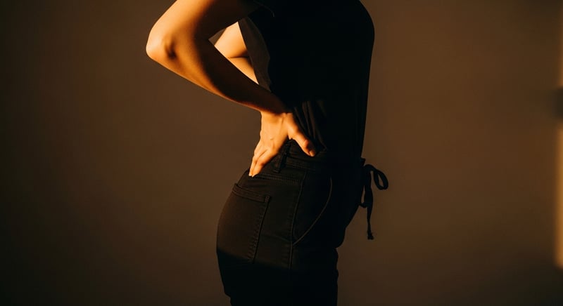 Editorial side-profile of a worker standing tall with hands on the low back easing into a gentle backward bend to reset between clients, warm amber light, no identifiable face
