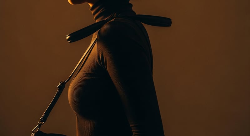 Side-profile editorial photograph of a woman walking with a bag strap on one shoulder, that shoulder lifted and rolled forward to keep the strap from sliding, face cropped above the nose