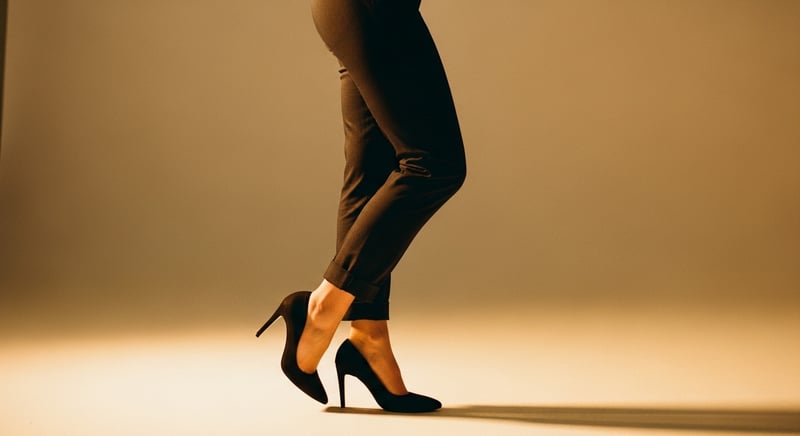 Side-profile editorial photograph of a person's lower body in high heels with weight tipped forward onto the balls of the feet, warm amber studio light