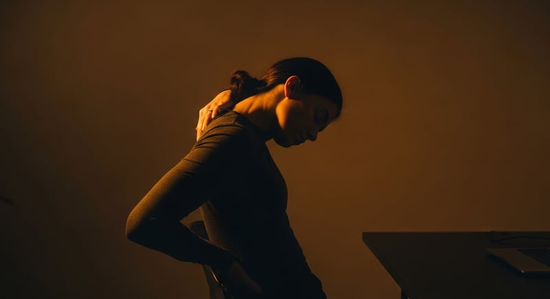 Editorial side-profile photograph of an anonymous person at a desk performing a seated neck stretch, head turned and chin dropped toward the armpit, warm amber side lighting, face cropped above the nose, no identifiable facial features