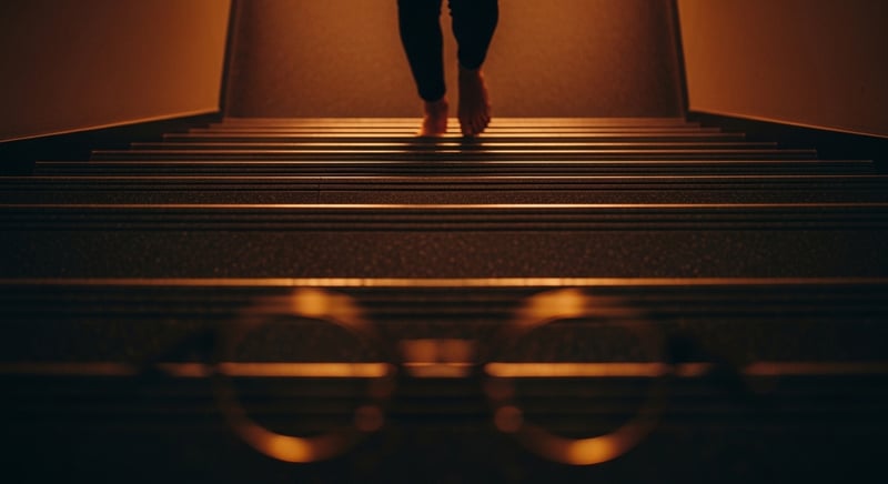 Editorial photograph looking down a flight of stairs past a foot mid-step, the lower steps softly blurred to suggest a compromised lower visual field, warm amber light