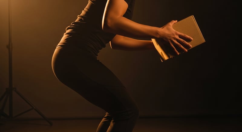 Editorial photograph of a person lifting a small light box with a visible jolt through the lower back, warm side lighting, face cropped above the nose